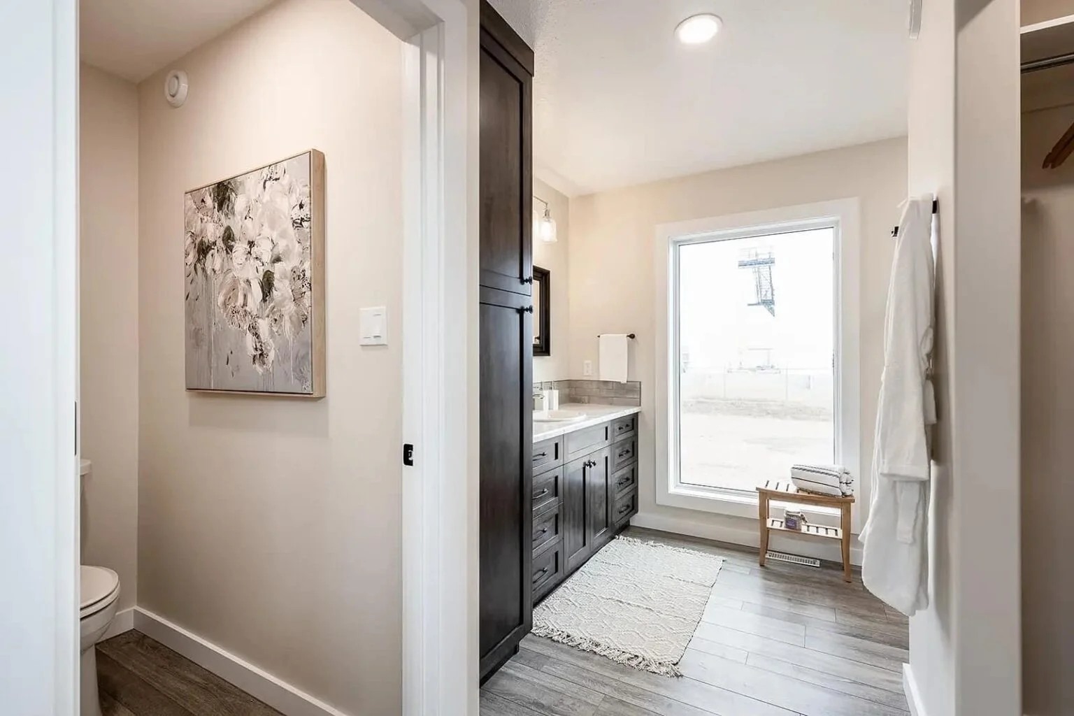 Modern bathroom with warm tones, featuring a dark wood vanity and large window. A soft rug and abstract painting add elegance and calmness.