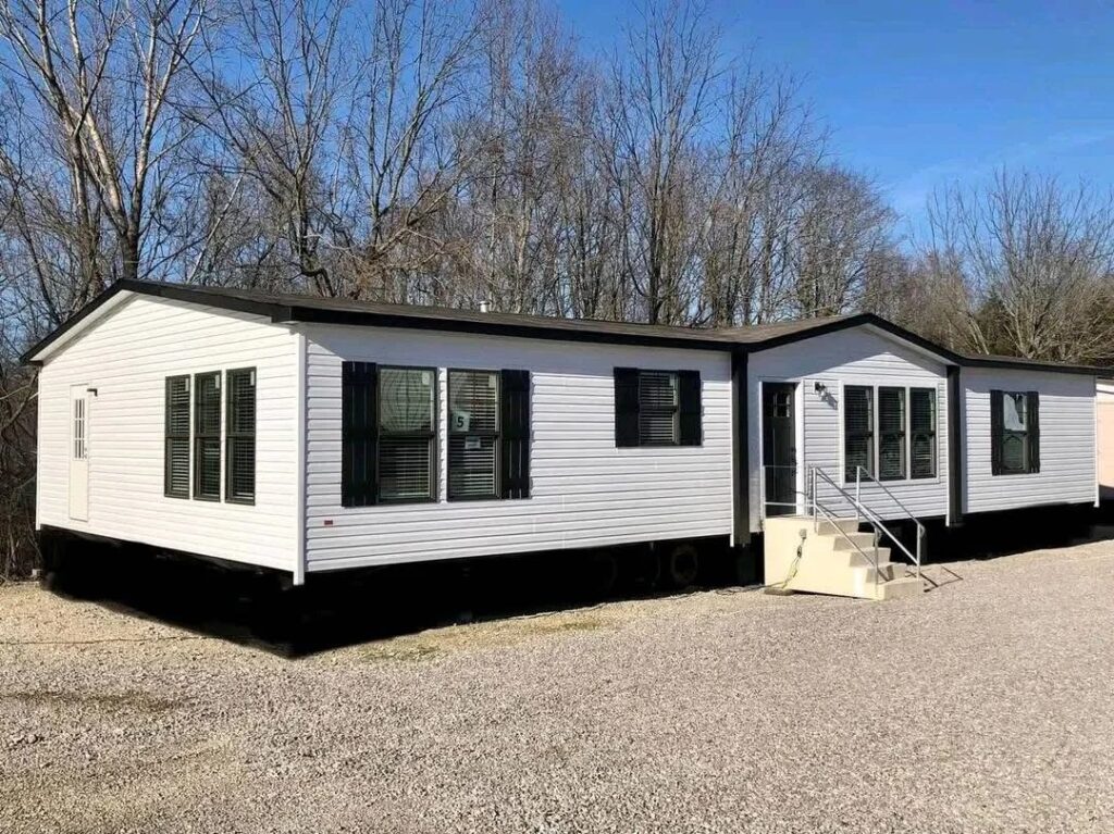 "A large white manufactured home with black windows and shutters, situated on a gravel lot. The house features white stairs leading to the entrance, with bare trees and a clear blue sky in the background."