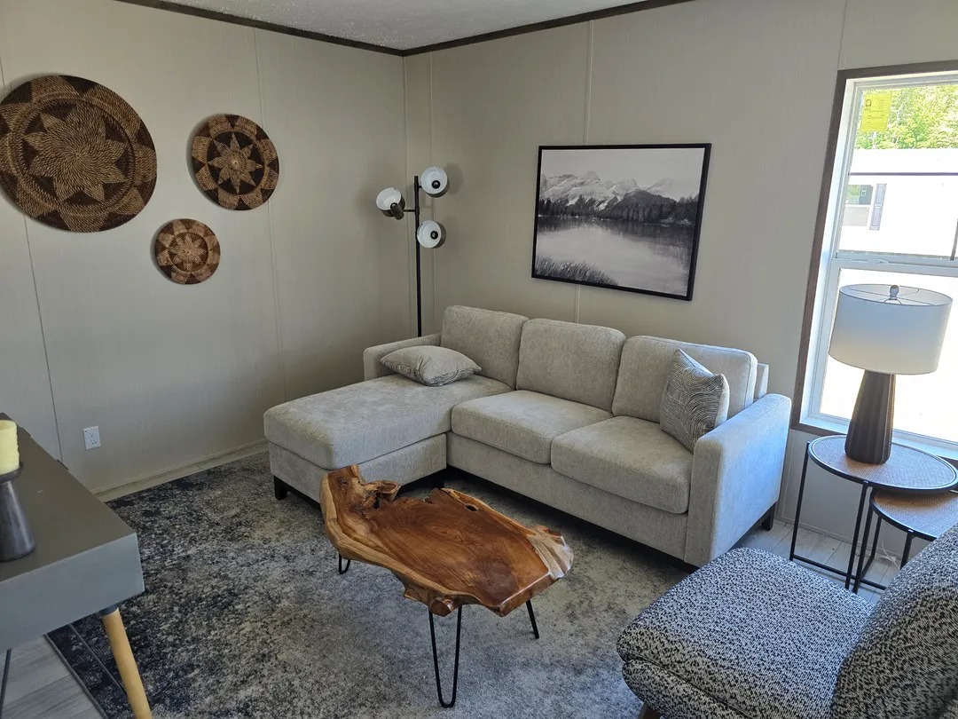 Cozy living room with light gray sectional sofa, rustic wood coffee table, and woven wall art. A floor lamp and a mountain landscape photo add warmth.