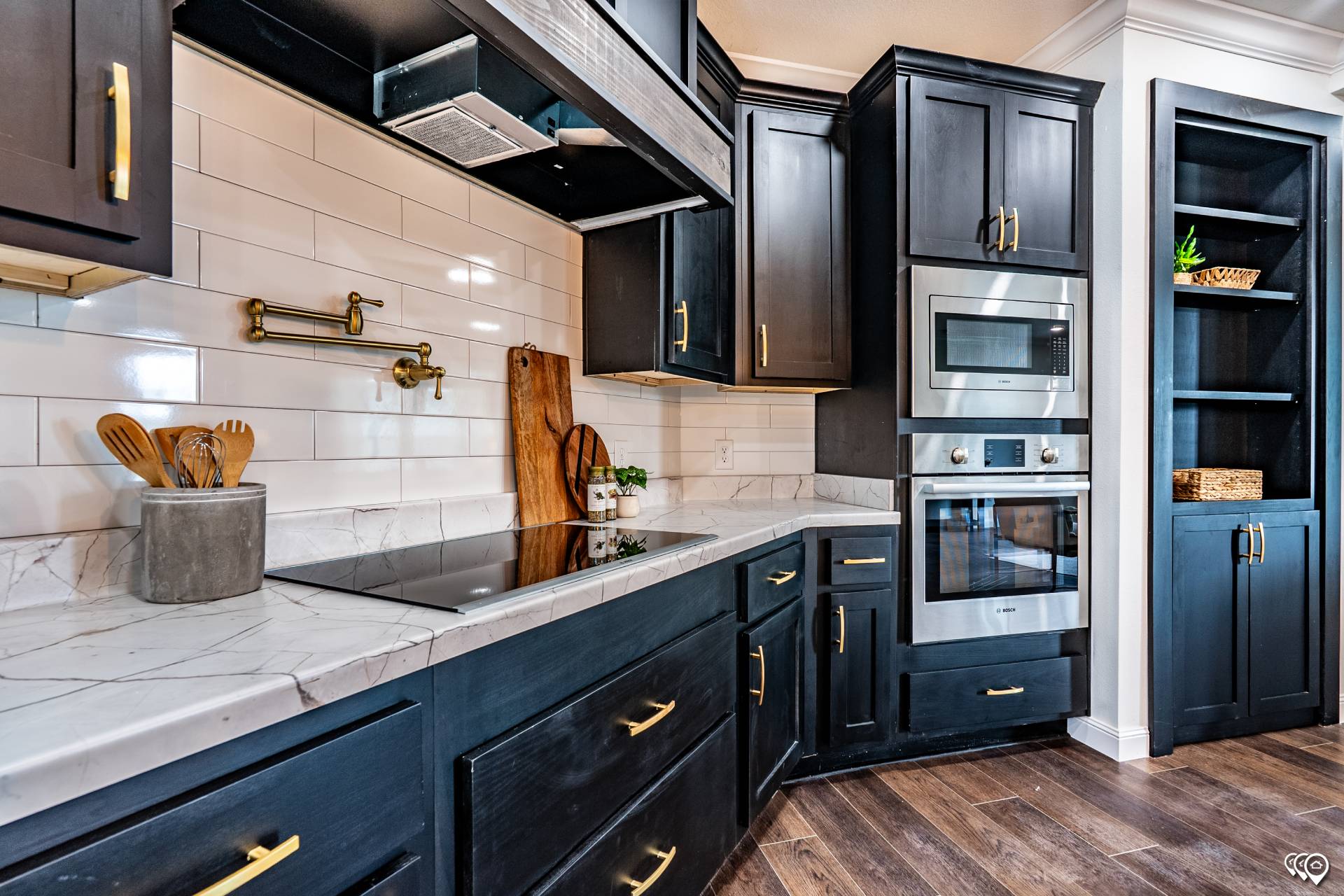 Modern kitchen with dark cabinets and gold handles, featuring a marble countertop, built-in oven and microwave, wooden floors, and white subway tiles.