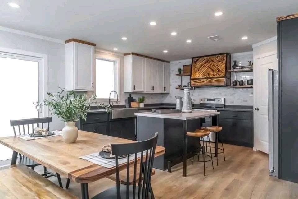 Modern kitchen with white and dark cabinets, wood accents, and gray countertops. A wooden dining table with black chairs and a vase adds warmth.