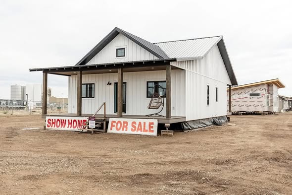 A small white show home with a gray roof and front porch is displayed for sale on a dirt lot. Large signs read "Show Home For Sale," creating an inviting and hopeful tone.