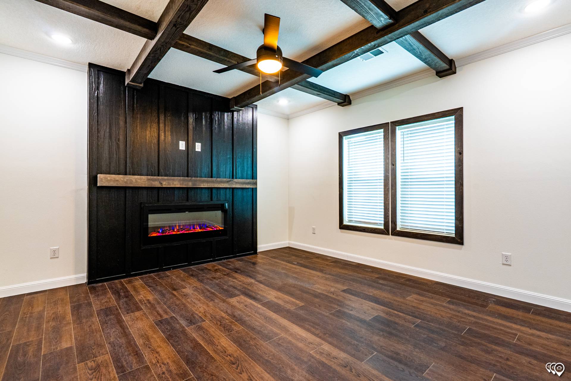 A modern living room with dark wood floors, a black accent wall featuring a sleek electric fireplace, exposed ceiling beams, and two large windows.