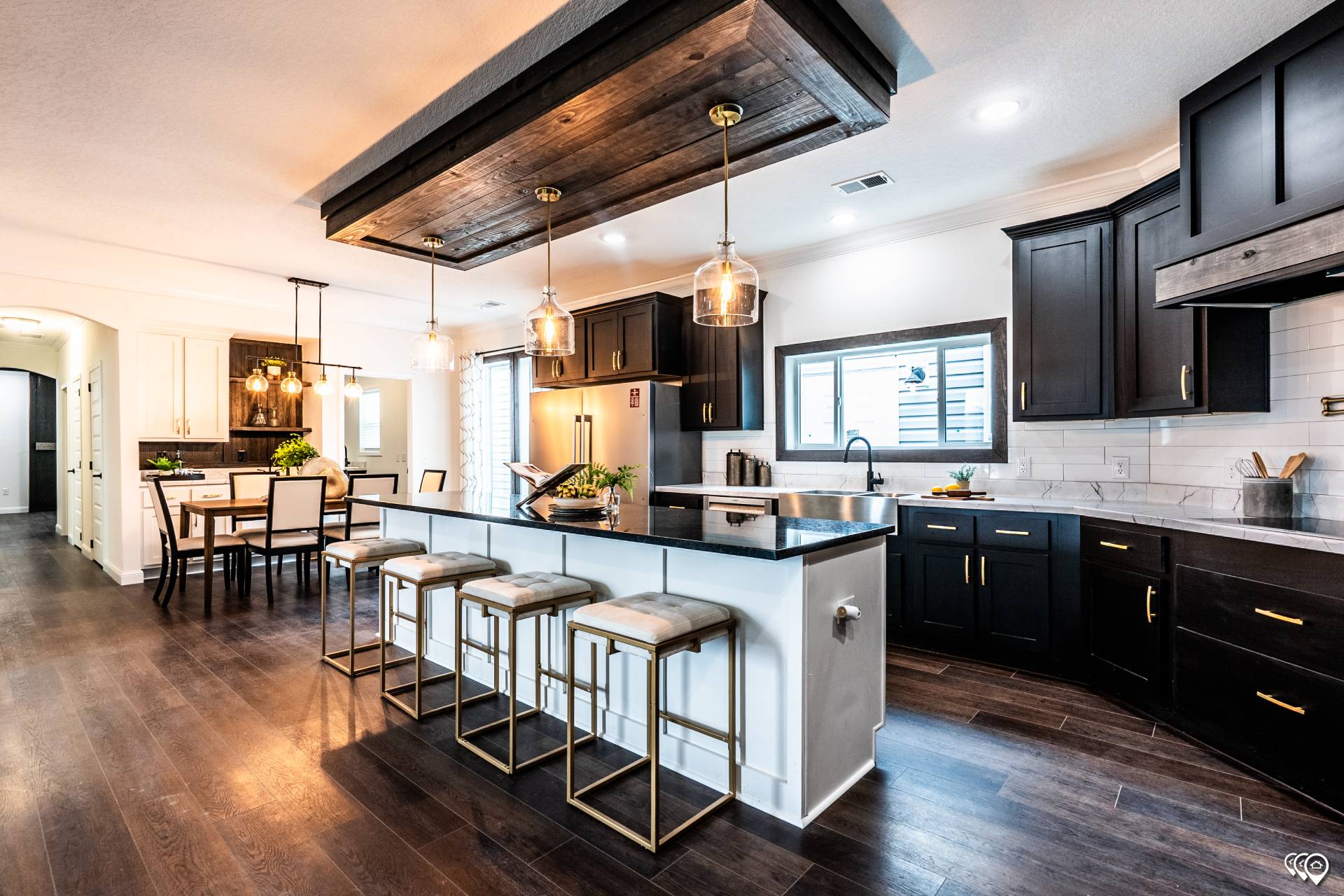 Modern kitchen and dining area with dark wood floors, black cabinets, and a large island. Three pendant lights hang above the island with four stools. The dining table is on the left, creating a warm and inviting space.