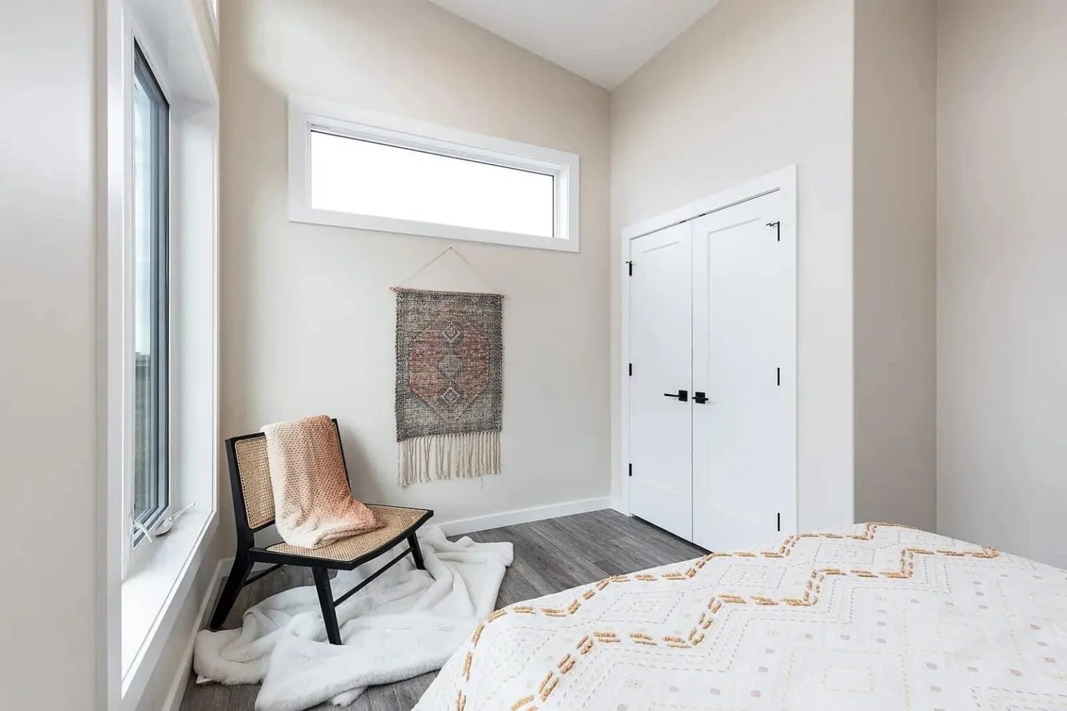 Minimalist bedroom with beige walls, featuring a window, wooden chair draped with a textured blanket, woven wall hanging, and a double closet door.