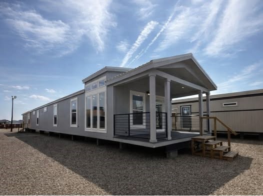 A modern gray modular home with a covered porch sits on a gravel lot under a blue sky with wispy clouds. Large windows and natural wood steps are prominent.