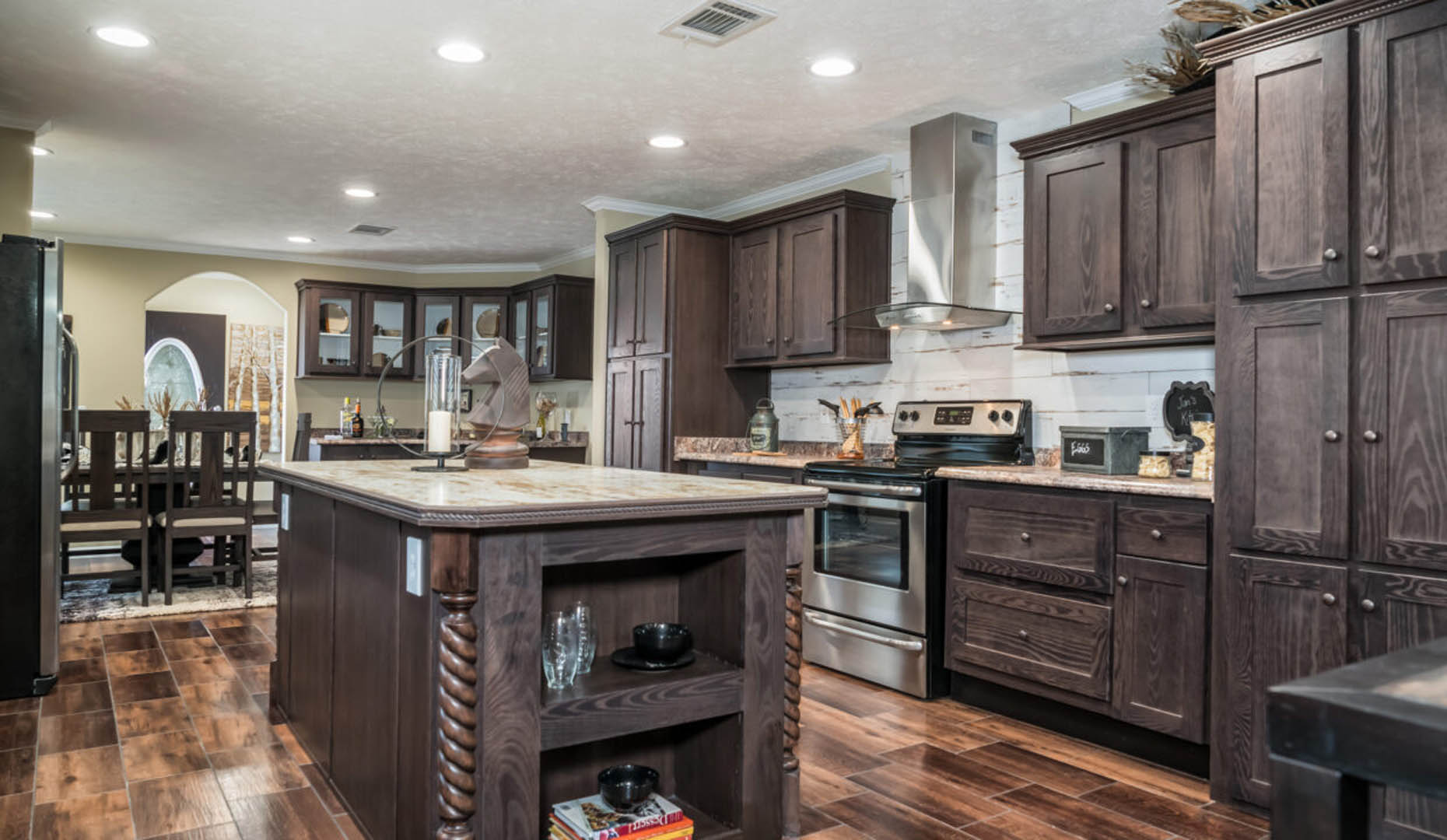 Spacious kitchen with dark wood cabinets, a large central island, stainless steel appliances, and wooden floors. Cozy, warm ambiance with soft lighting.