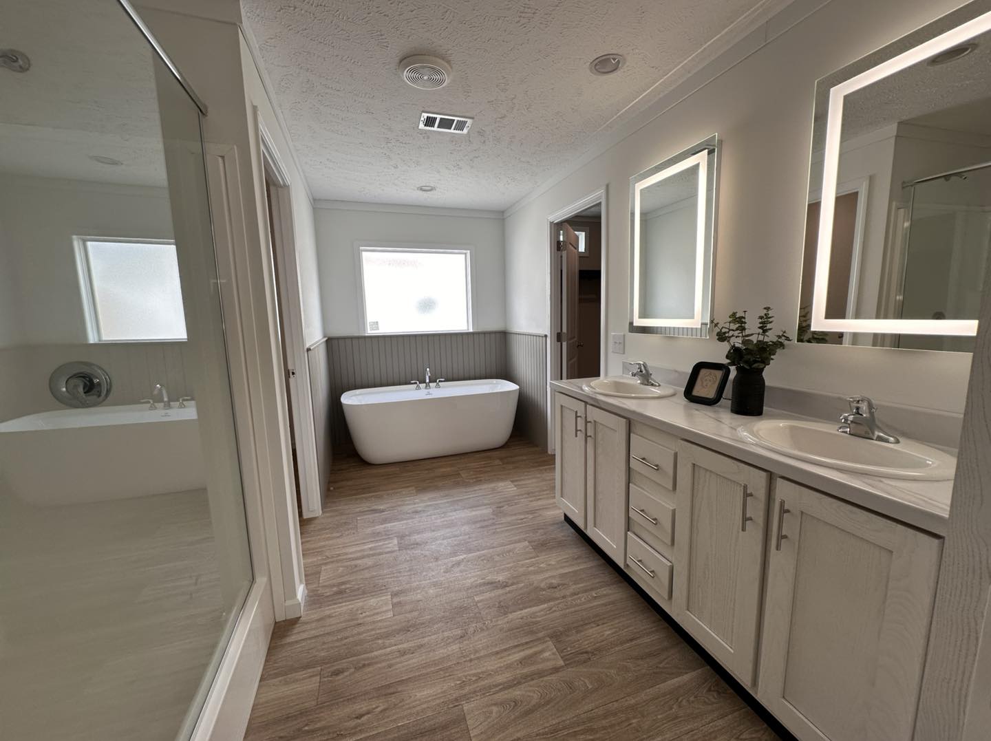 Luxury high-end manufactured home showcase featuring expansive double-wide floor plans with dark charcoal horizontal siding and rustic wood shutters. Modern interiors highlight a spacious open-concept living area with an integrated electric fireplace and shiplap accent wall, a gourmet kitchen with a large marble-look island and herringbone tile backsplash, and custom floor-to-ceiling pantry storage with integrated pull-out drawers. The design features vaulted shiplap ceilings with dark wood beams, cozy bedrooms with modern iron or wood frames, a stylish master suite with a black accent wall and cowhide rug, a master bathroom with a freestanding soaking tub and LED-mirrored double vanity, and a tranquil covered front porch with a dark wood-plank ceiling and a hanging wicker egg chair.