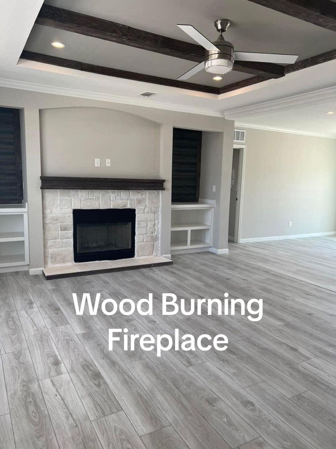 Modern living room with light wood flooring, a stone fireplace, built-in shelves, a ceiling fan, and dark wooden beam accents on the ceiling.