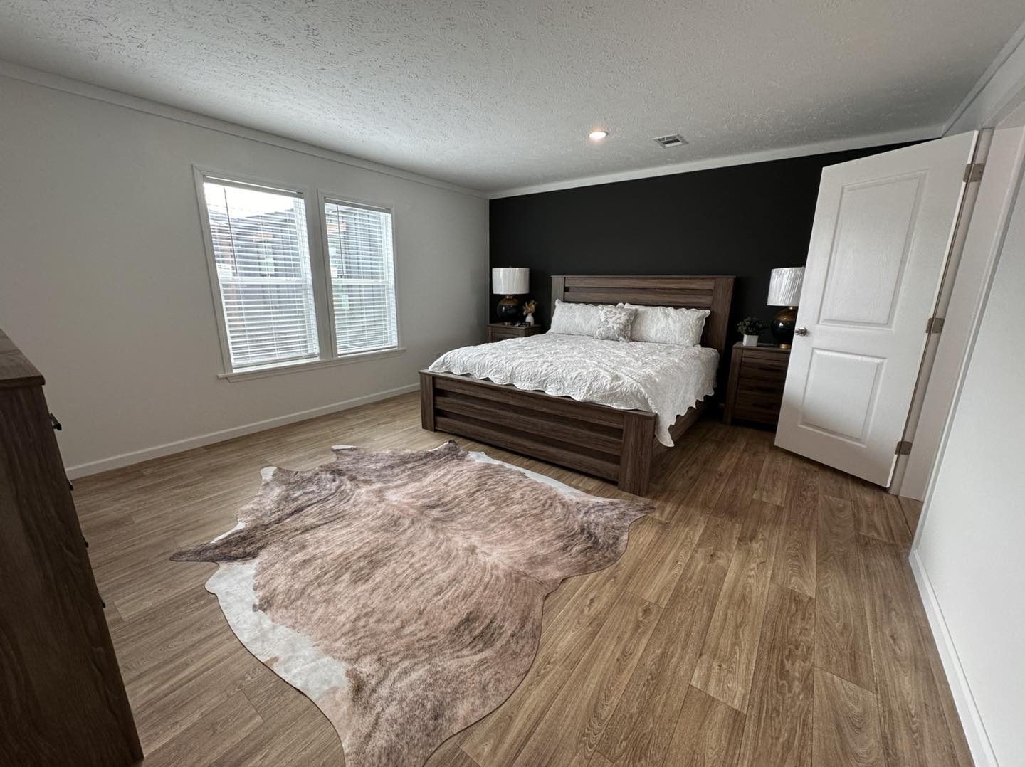 High-end manufactured home showcase featuring expansive double-wide models with dark charcoal horizontal siding and rustic wood shutters. Luxury interiors highlight a spacious open-concept living area with a grey electric fireplace and shiplap accent wall, a gourmet kitchen with a large marble-look island and herringbone tile backsplash, and custom floor-to-ceiling pantry storage with integrated pull-out drawers. The design is completed by vaulted shiplap ceilings with dark wood beams, cozy bedrooms with modern iron or wood frames, a stylish master suite with a black accent wall and cowhide rug, and a tranquil covered front porch with a dark wood-plank ceiling and a hanging wicker egg chair.