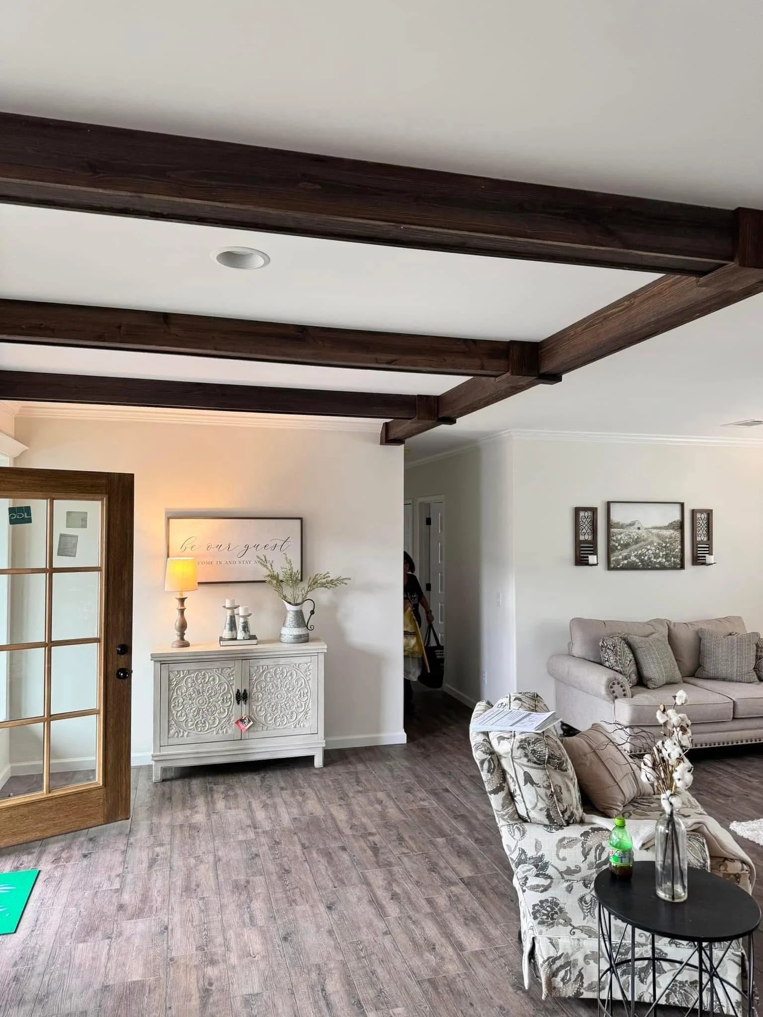 A cozy living room featuring wooden ceiling beams and laminate flooring. A gray couch with floral pillows sits near a small round table. A cabinet with decorative items and a lamp adds warmth to the space.