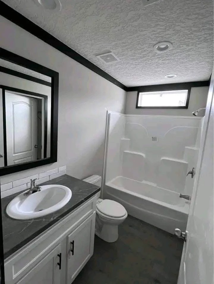 Small, modern bathroom with white toilet, sink, and bathtub. Dark countertop and trim contrast with light walls and ceiling. Bright, minimal, and clean.