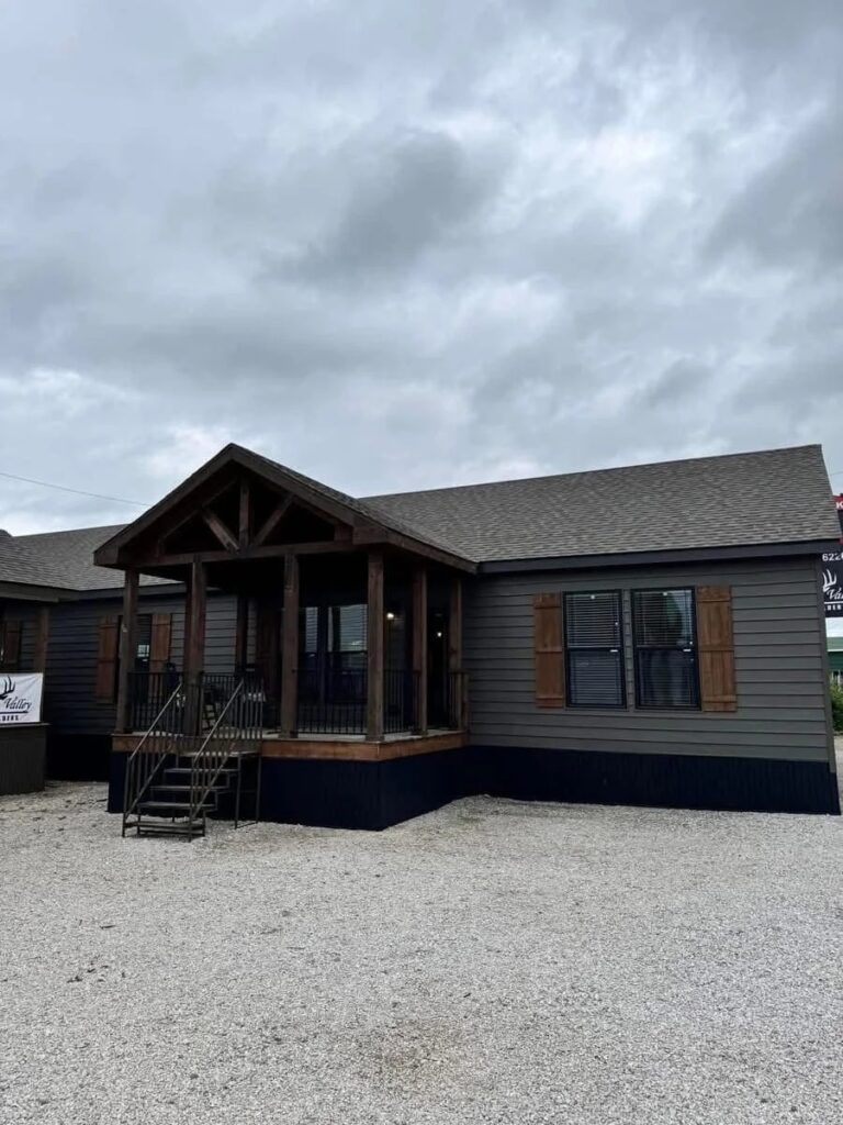 A modern, dark gray prefab home under a cloudy sky. It features a wooden porch with stairs, large windows with shutters, and a gravel driveway, creating a cozy yet subdued ambiance.