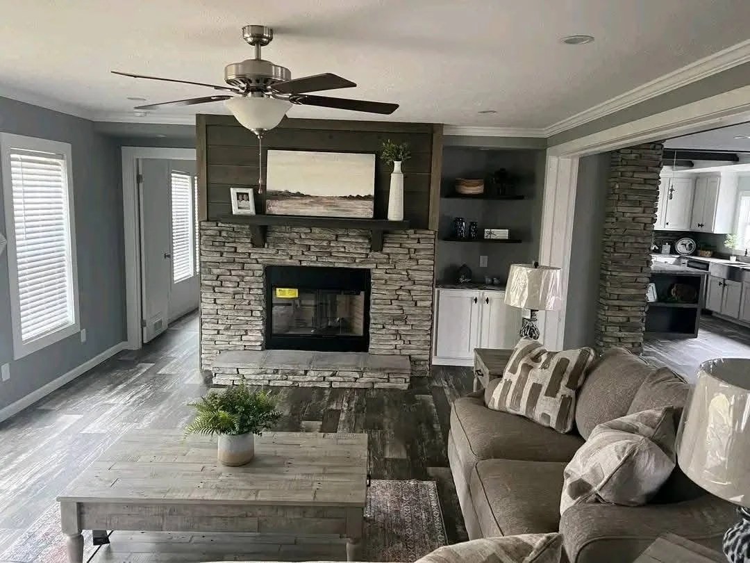 Cozy living room with a stone fireplace, neutral tones, and a ceiling fan. A gray couch with patterned pillows faces a rustic coffee table.