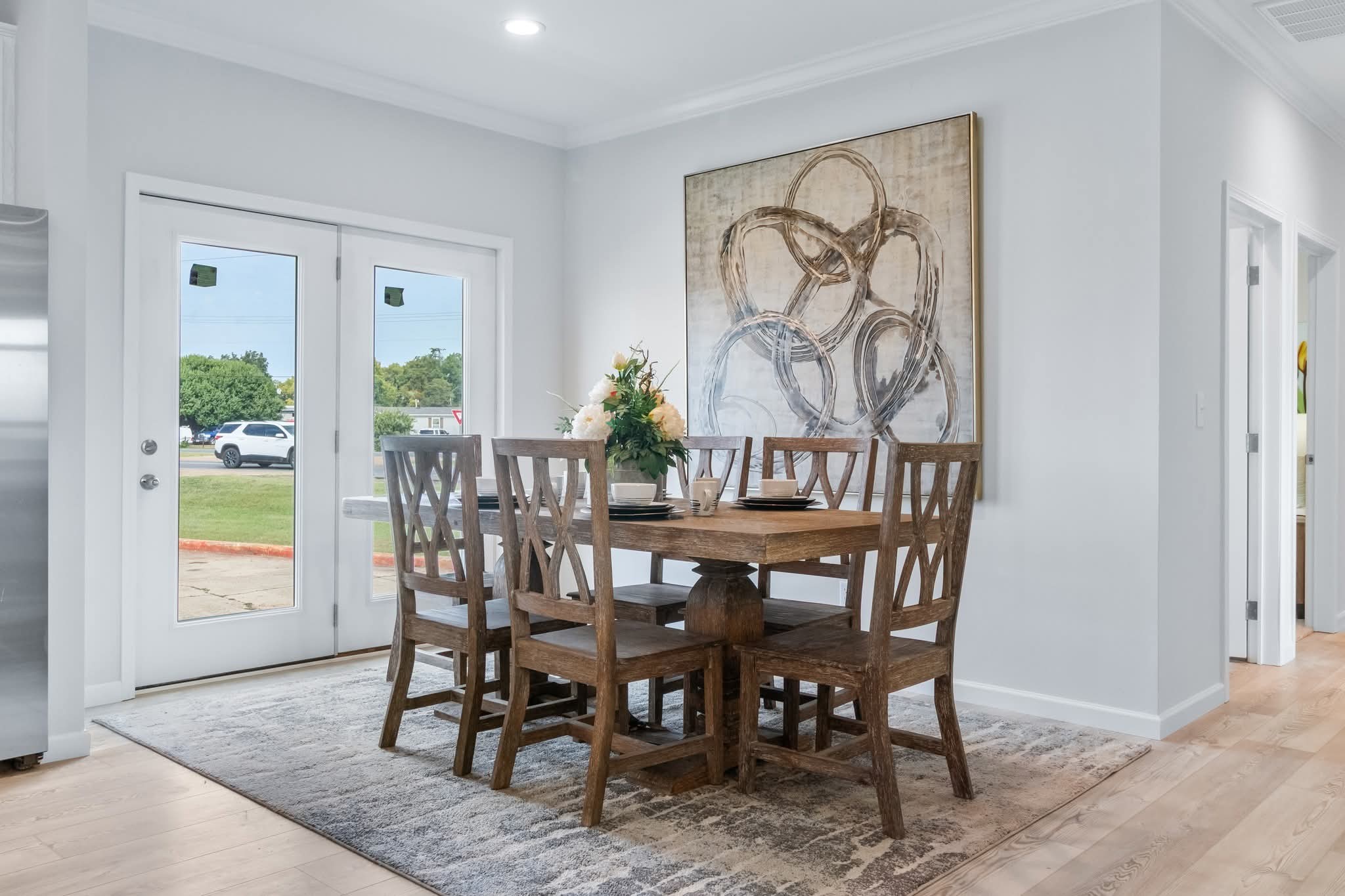Dining room with a wooden table and six chairs on a patterned rug. Large abstract art hangs on a white wall, and light pours in from glass doors.