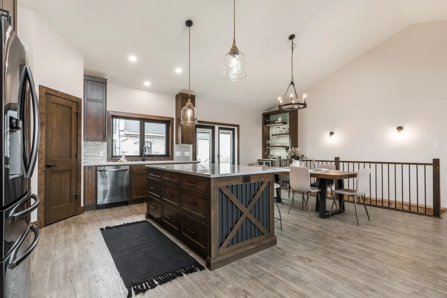 An open-concept kitchen and dining area with a modern rustic feel. A large dark wood kitchen island with an 'X' crossbeam detail and a granite countertop sits in the center, lit by two glass pendant lights. In the background, a dining table with light grey chairs is positioned under a black circular chandelier next to a staircase with a black metal railing. The space features dark wood cabinets, light grey flooring, and white walls.