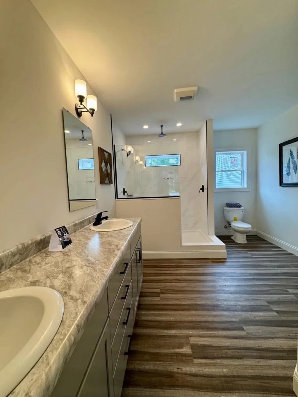 Modern bathroom with marble countertops, dual sinks, and bright lighting. Features a glass shower, wood flooring, and artwork on beige walls.