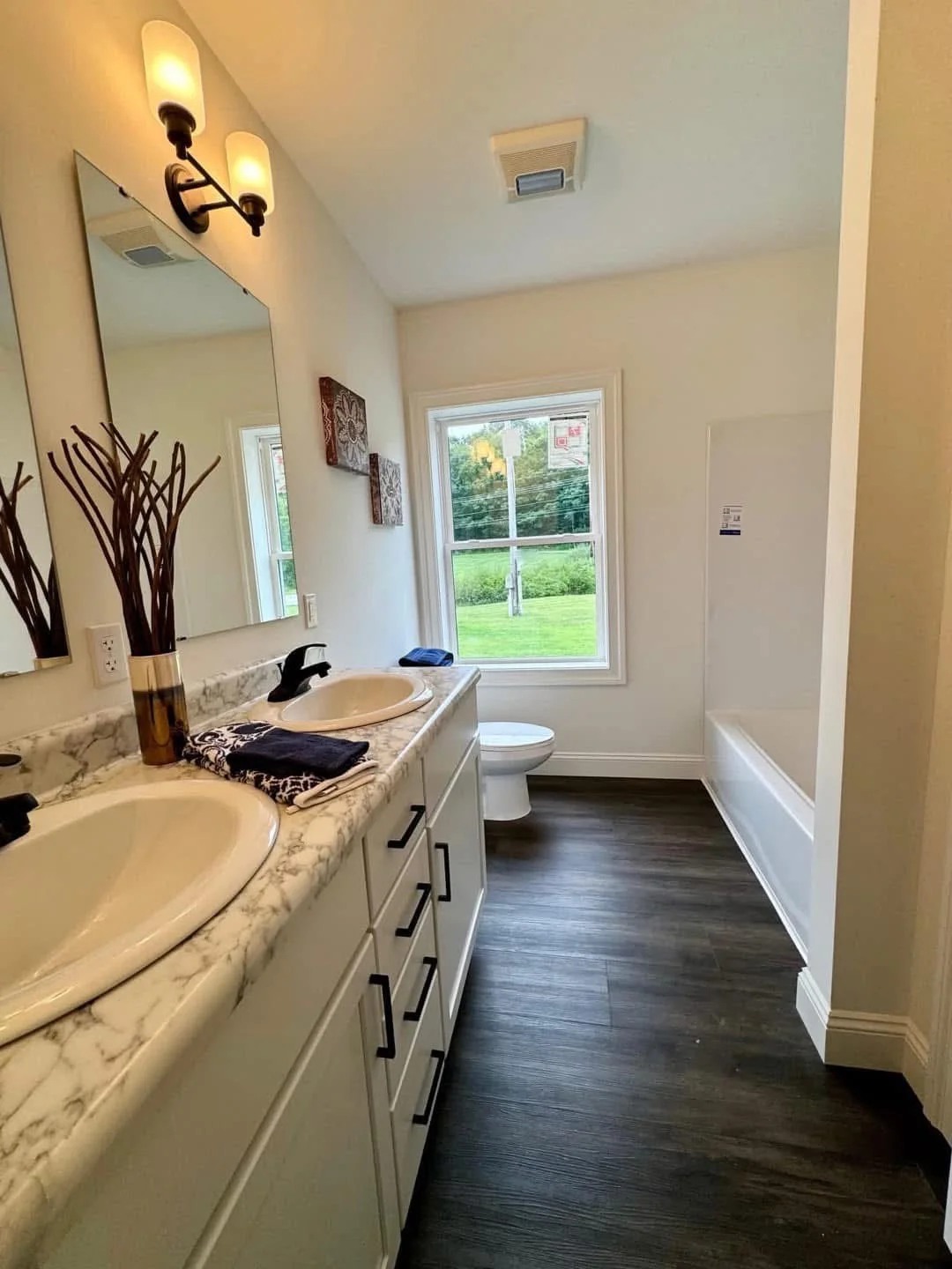 Modern bathroom with dual marble sinks, dark faucets, and decorative vases. Wood floor leads to a tub. A window shows lush greenery outside.