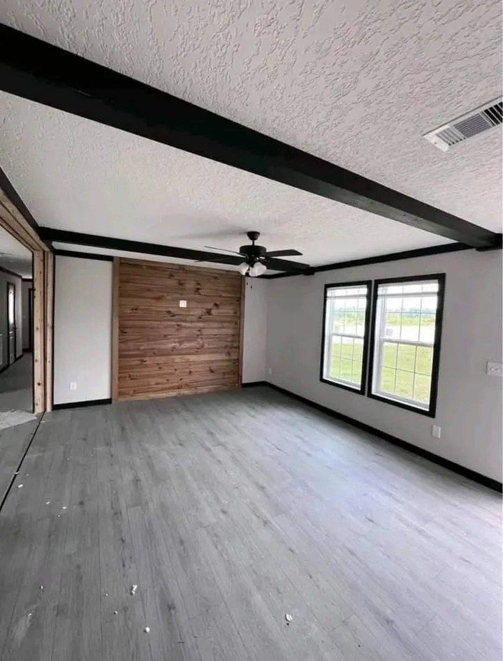 Spacious living room with light gray flooring, textured white ceiling, and dark wood beams. A ceiling fan is centered above a wood accent wall, and bright natural light streams through two large windows. The room feels modern and airy.