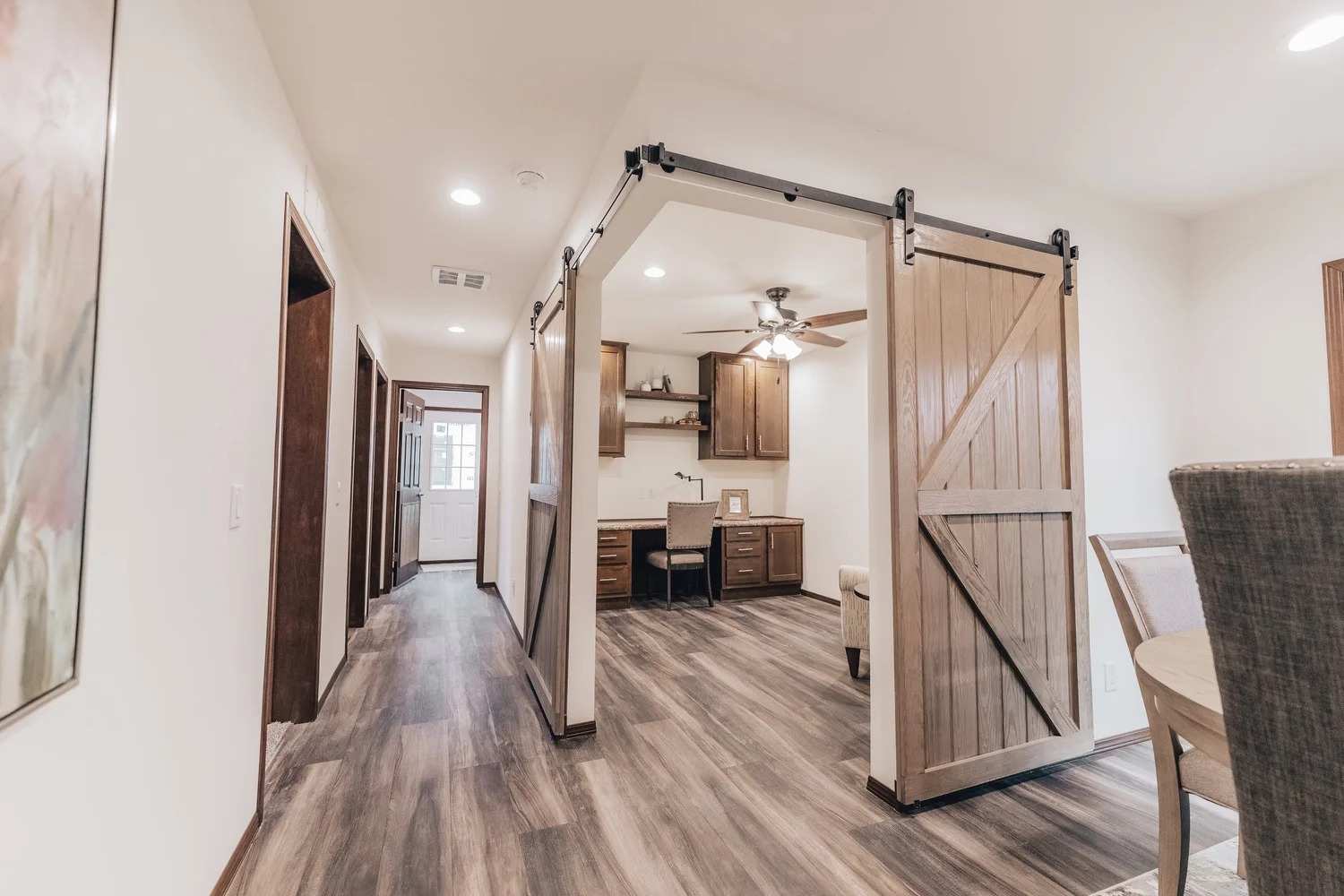Warmly lit hallway with wood flooring leads to a cozy office space shown through open barn doors. A dining area is partially visible on the right.
