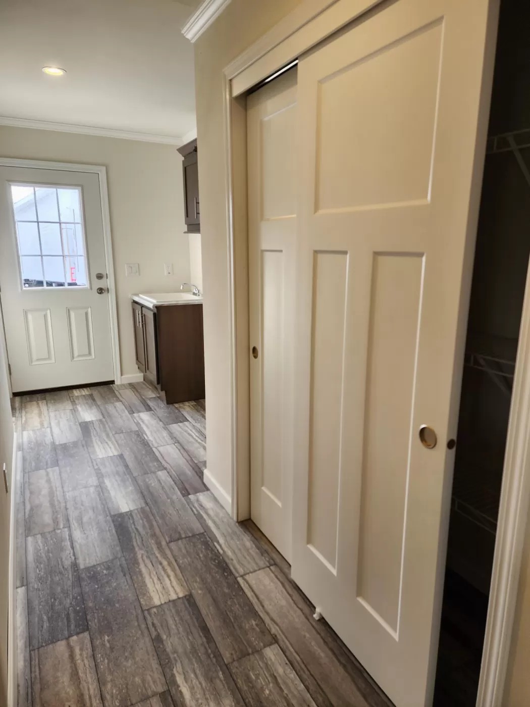 Hallway with a sliding closet door on the right and a light wood tiled floor. At the end, a door with a window panel leads outside. The space feels bright and clean.