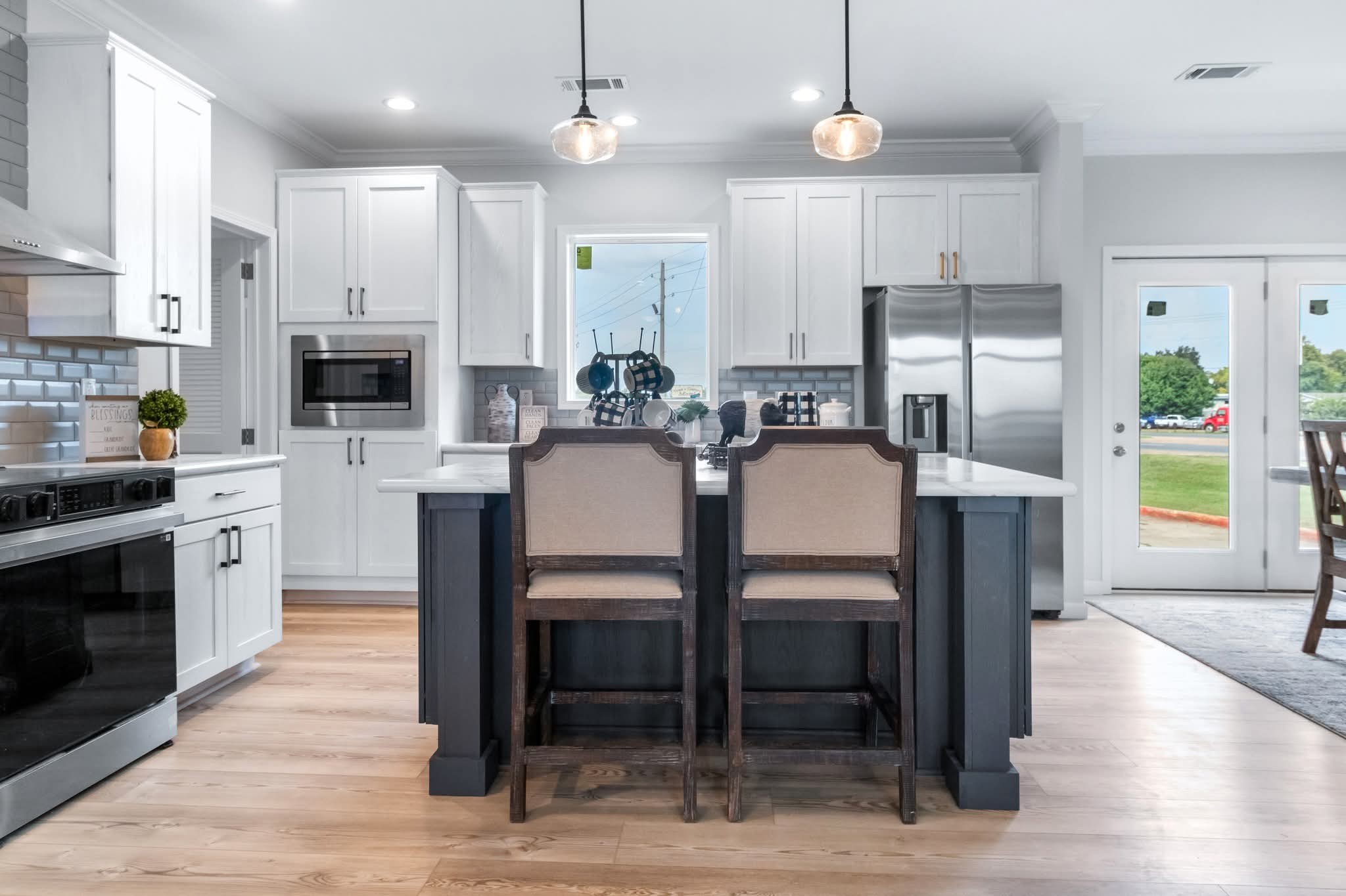 Modern kitchen with white cabinets, stainless steel appliances, and a central island with two cushioned stools. Bright and airy with natural light.