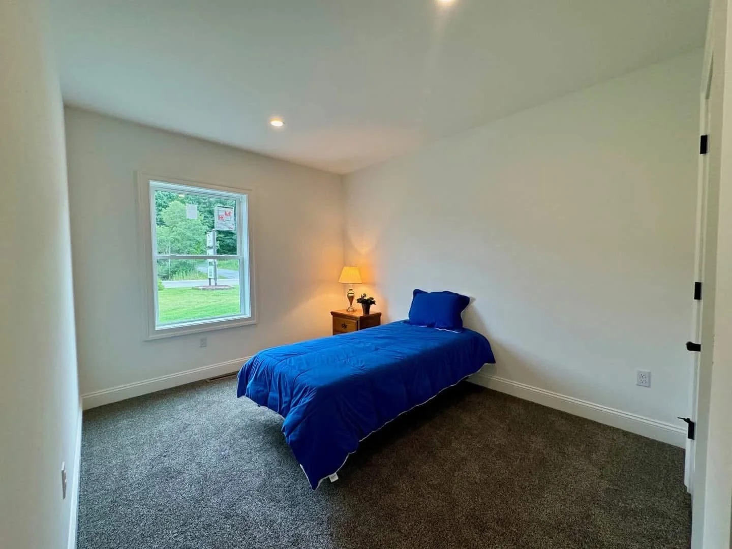 A small, bright bedroom with a single bed covered in a royal blue comforter and pillow. A lit lamp on a wooden nightstand adds warmth beside the window.