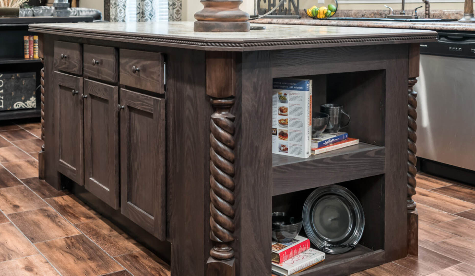 Rustic kitchen island with dark wood, intricate twisted columns, cabinets, and open shelves holding books and dishes, on a wooden floor. Cozy ambiance.