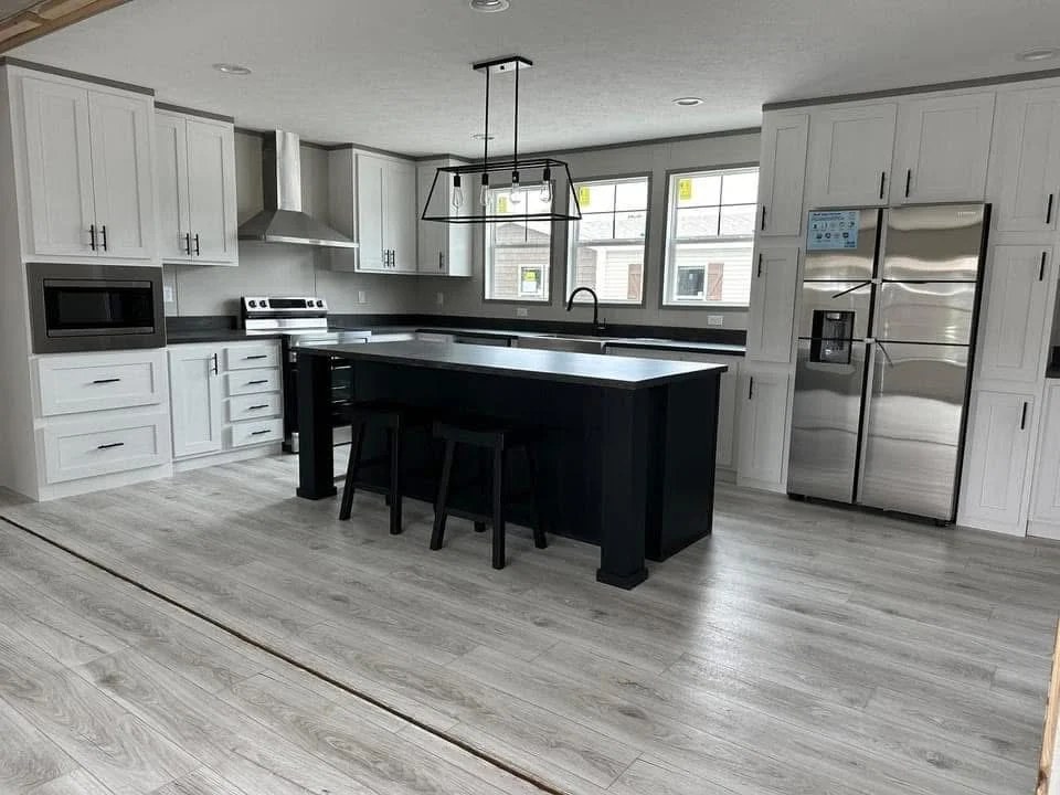 Modern kitchen with light wood floors, white cabinets, and a dark island. Features stainless steel appliances and large windows, creating a bright, sleek atmosphere.