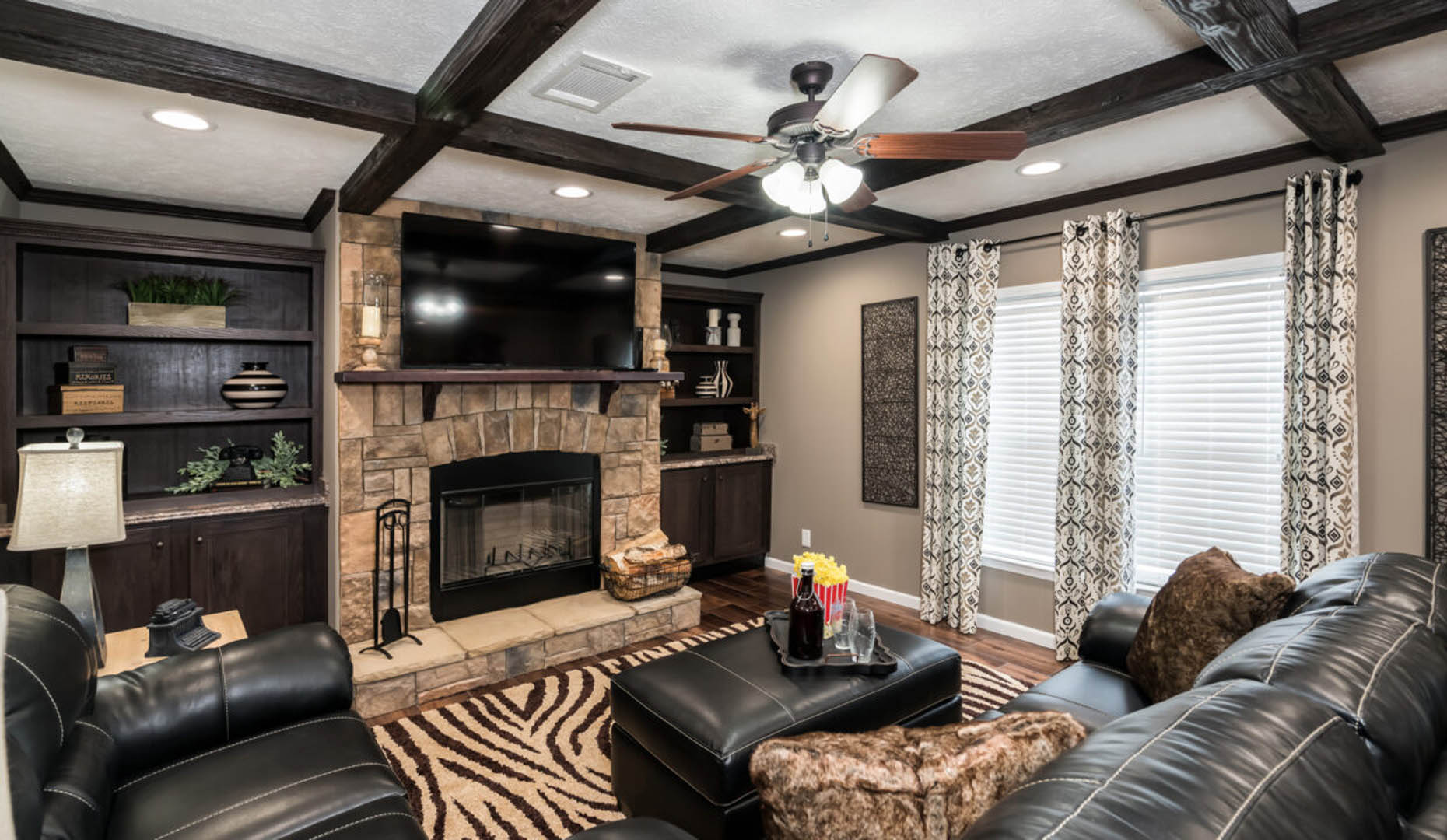 Cozy living room with black leather sofas, stone fireplace, and a TV. Zebra-patterned rug adds flair, while large windows and shelves create a warm, welcoming vibe.