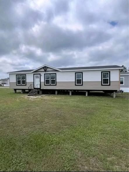 Single-story modular home on stilts against a cloudy sky, with a few stairs leading to the entrance. It sits on a grassy field, conveying simplicity.