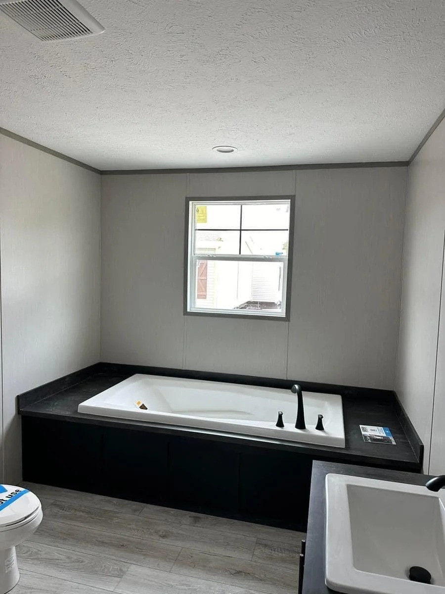 Minimalist bathroom with a large rectangular tub beneath a window. The room features light gray walls, wood-look flooring, and a white sink.