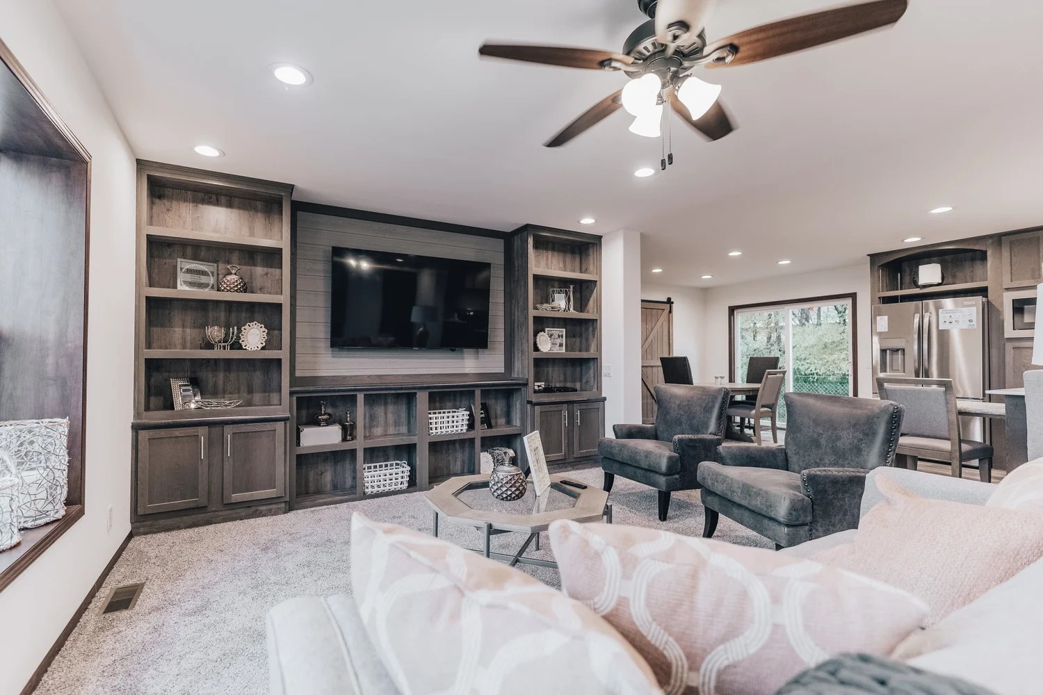 Spacious living room with modern decor, featuring a ceiling fan, built-in shelves, a large TV, plush armchairs, and a glimpse of the dining area. Cozy and inviting ambiance.