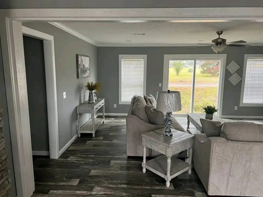 Modern living room with gray walls and flooring, featuring a beige sofa and armchair. Large window, ceiling fan, and decorative plants create a cozy atmosphere.