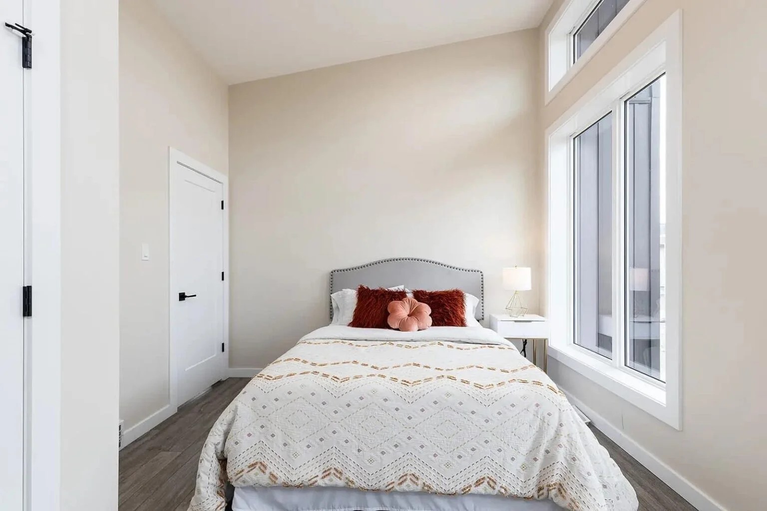 Bright bedroom with a bed featuring a white and gold patterned duvet, red and pink pillows, white nightstand, lamp, and large windows. Calm and inviting.