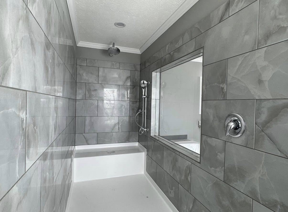 Modern bathroom with sleek gray tiles and a wall mirror. Features a rain showerhead, handheld shower, and a rectangular white bathtub. Clean and minimalist design.