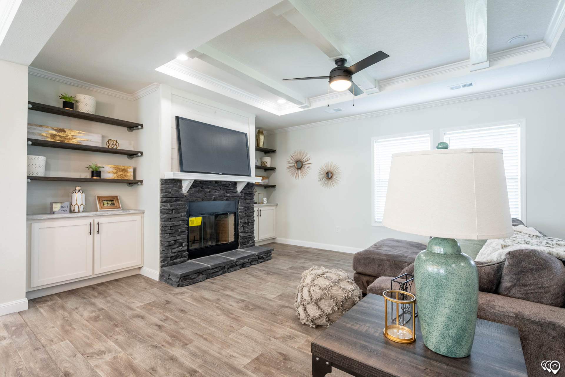 Living room with a modern, cozy feel. Features a stone fireplace, mounted TV, white cabinets, wood shelves, and a soft gray sectional. Accents include a teal lamp, decorative pillows, and a geometric pouf.