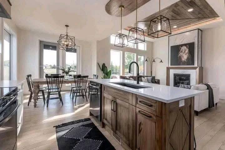 Modern kitchen and dining area with wooden cabinets, a white island, and geometric pendant lights. Sunlit space with a cozy living area and fireplace.