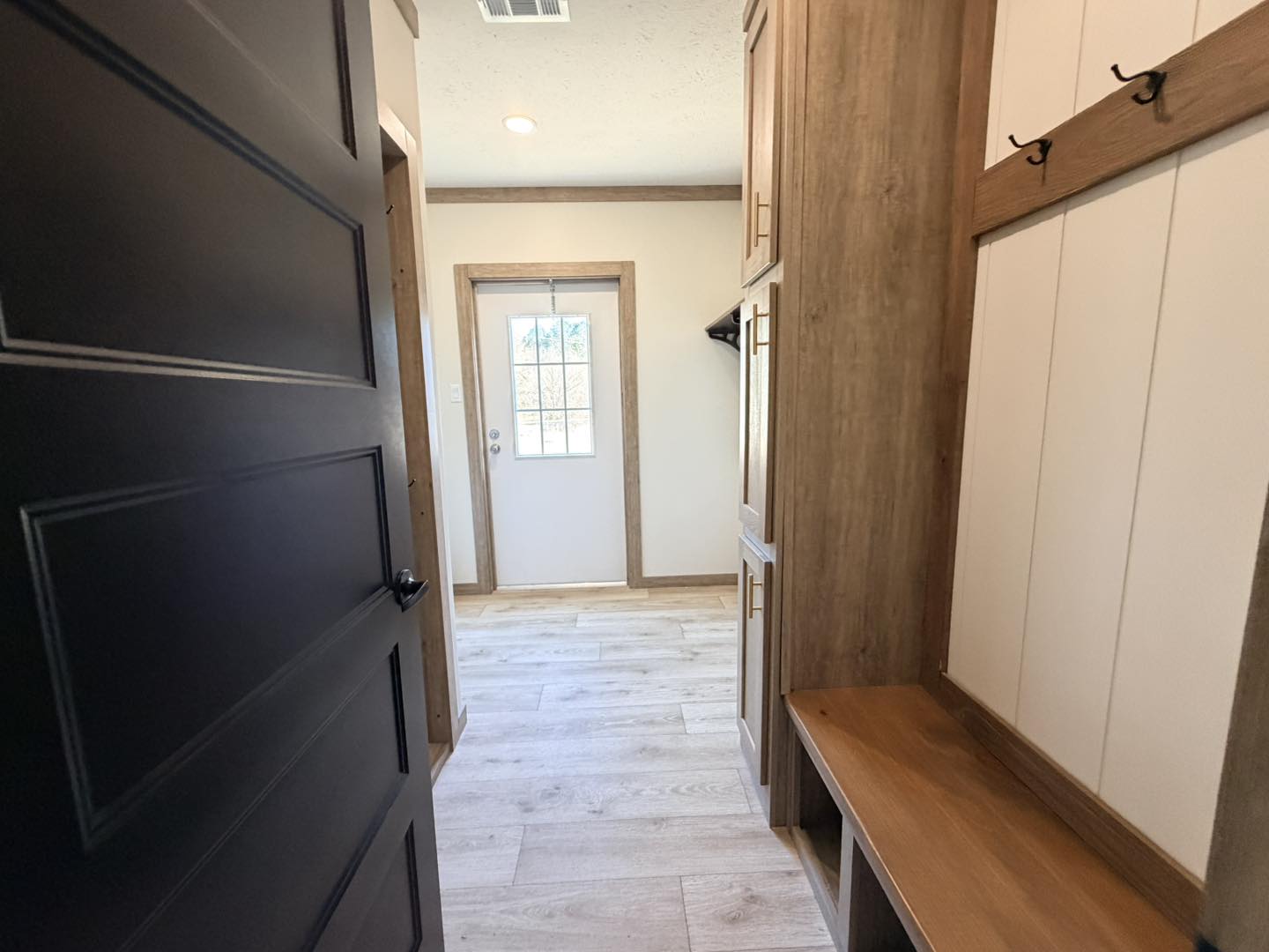 Open black door leading to a bright mudroom with light wood floors, a white wall, wood accents, and hooks. Sunlight streams through a small window.