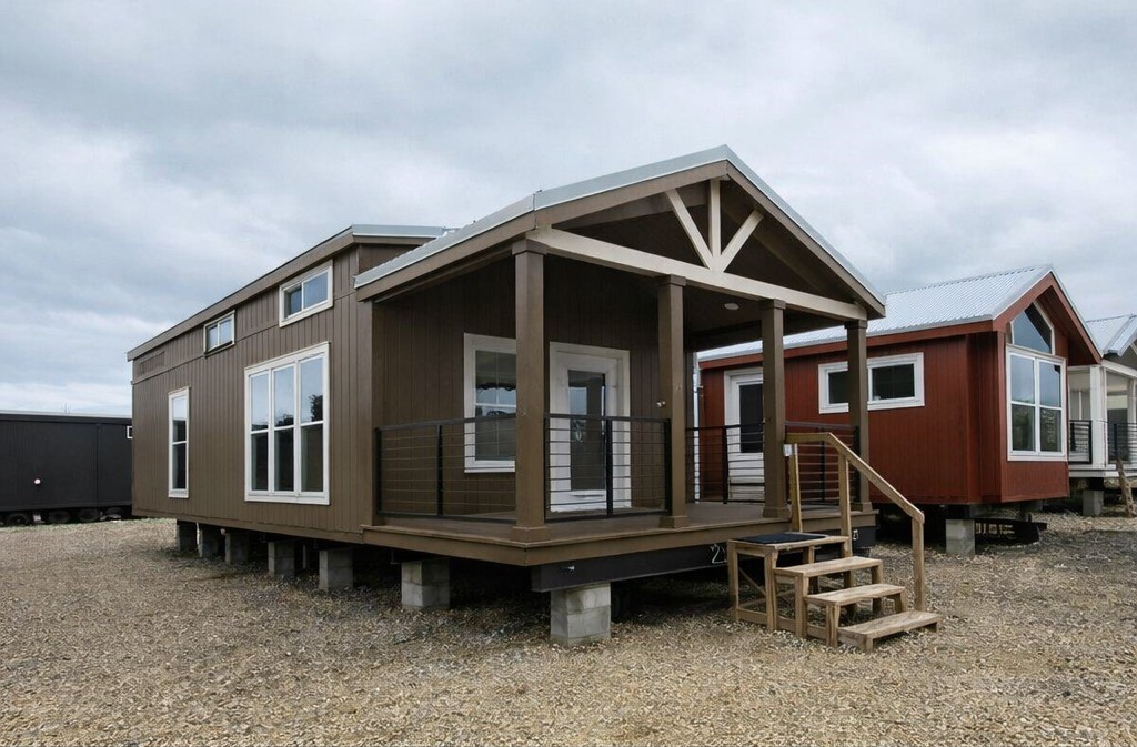 A brown tiny house with a gabled roof and elevated porch on gravel. Simple steps lead to the entrance. Overcast sky suggests a calm, modern rural setting.