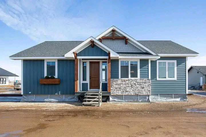 A modern single-story house with blue siding, stone accents, and a wooden front door. It sits on a dirt lot under a clear blue sky, conveying a fresh, inviting feel.