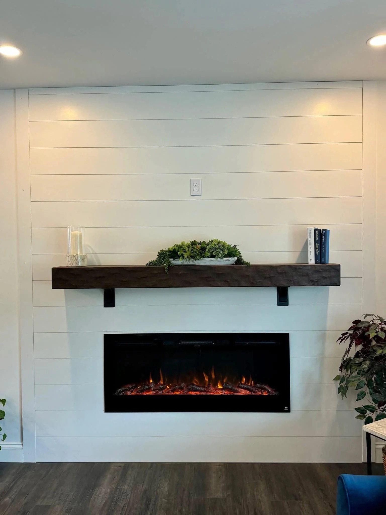 Electric fireplace with glowing embers set in a white shiplap wall. Above, a dark wooden mantel holds greenery, candles, and books, creating a cozy atmosphere.