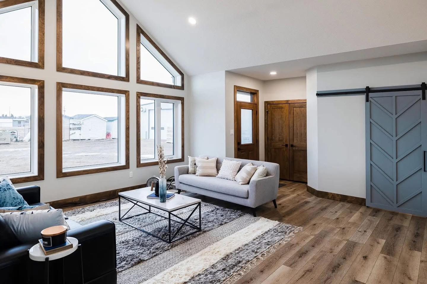 A comprehensive real estate gallery showcasing a 1,856 sq. ft. modern farmhouse-style home with 3 bedrooms and 2.5 bathrooms. The exterior features light grey horizontal siding with dark trim, a gabled roofline, and large vaulted windows. Interior highlights include an open-concept living room with vaulted ceilings, dark wood beams, a massive floor-to-ceiling stone fireplace, and contemporary furniture. The kitchen is designed with grey cabinetry, a large marbled island with three cushioned barstools, an industrial Edison-bulb chandelier, and a white farmhouse sink. Additional spaces shown include a dining area with a dark wood table and forest-themed wall art, a master bathroom with a freestanding soaking tub and a marble-tiled walk-in shower, and various entryways featuring sliding barn doors and built-in decorative shelving.