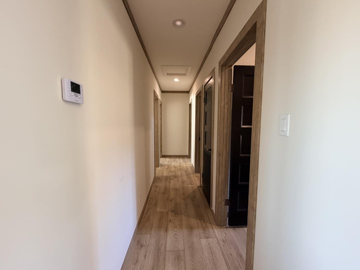 A bright, narrow hallway with light wood flooring and cream walls. Three doors line the right side, and a white thermostat is on the left wall.