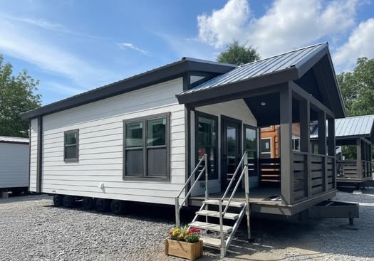 Modern tiny house with white siding and a dark roof. A small porch with plants in front adds charm. The sky is blue with a few clouds.