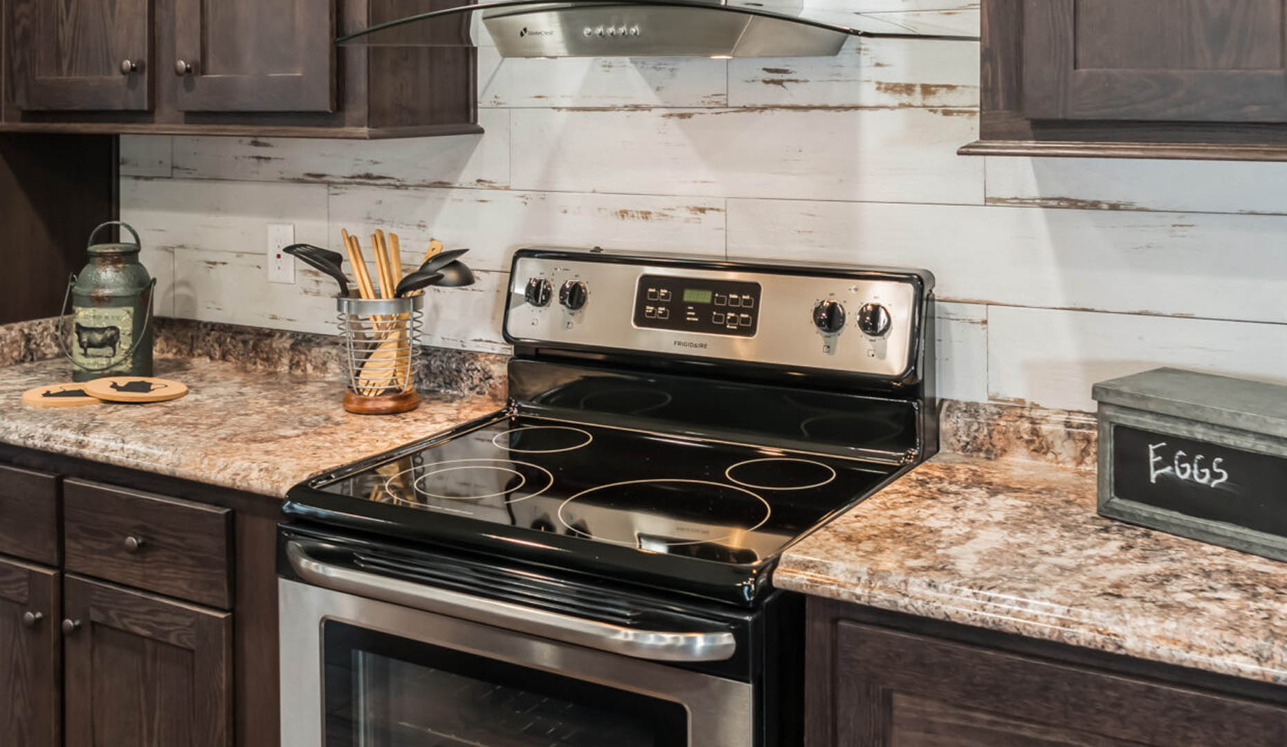 Modern kitchen with a stainless steel electric stove, dark wood cabinets, and a rustic backsplash. Counter holds a utensil jar and vintage decor.