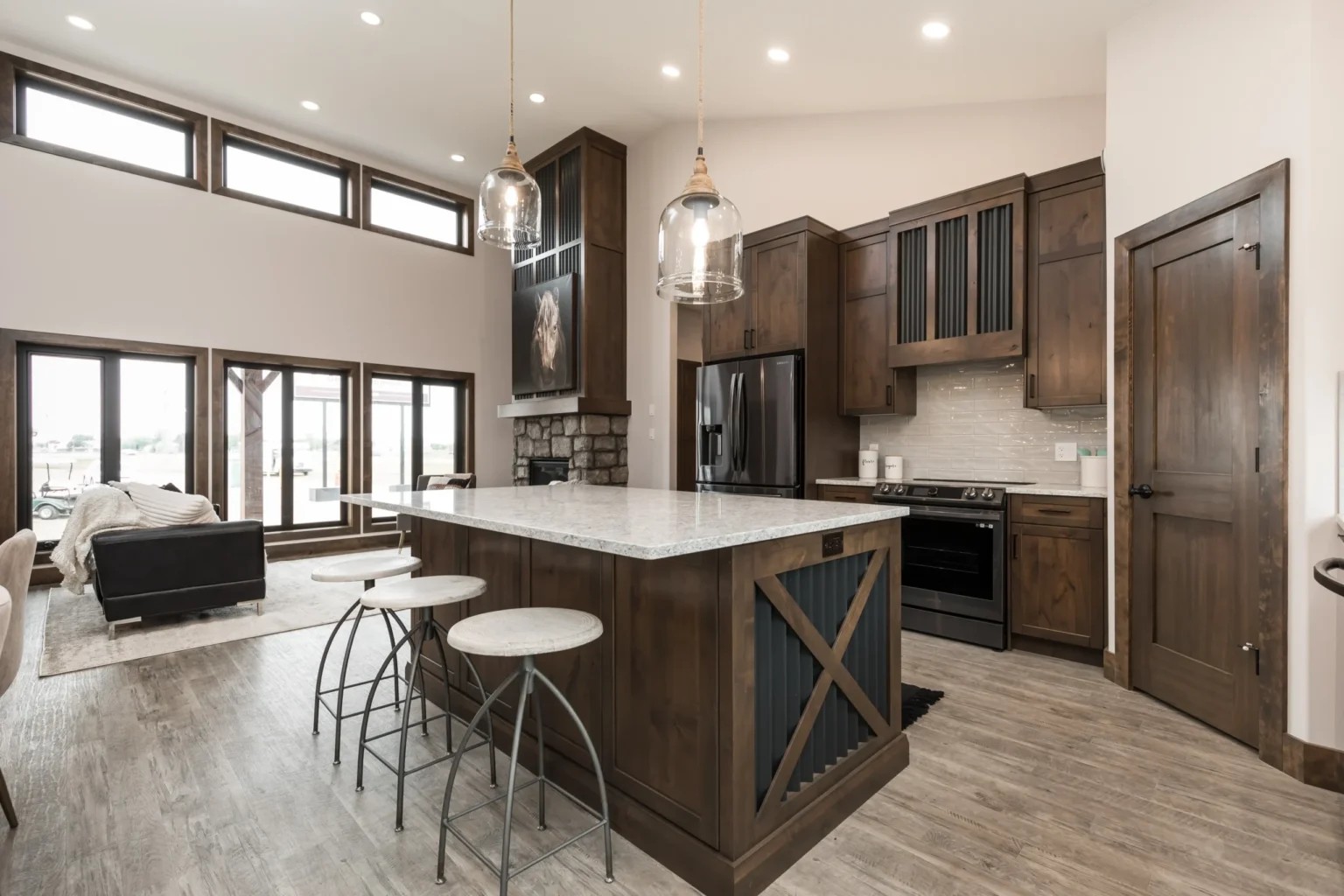 "A rustic modern open-concept kitchen with dark wood cabinets, a large center island with a white countertop, and industrial bar stools. The room features black stainless steel appliances, glass pendant lights, and light grey wood-look flooring. Large black-framed windows and a stone fireplace are visible in the background living area."