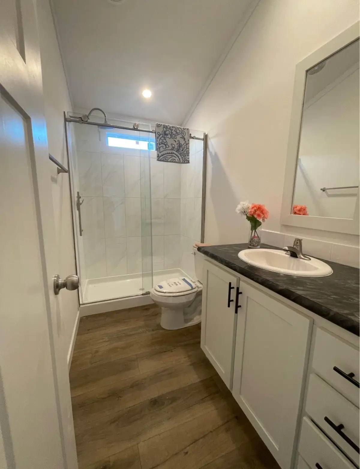 A small, bright bathroom features a tiled shower, wooden flooring, and a white vanity with a black countertop. A vase with flowers adds a touch of color.