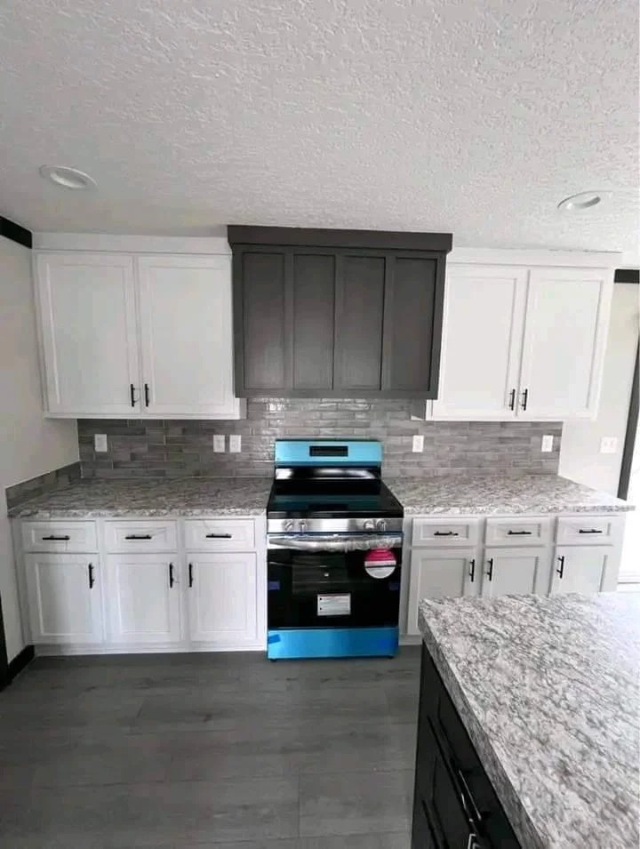 Modern kitchen with a stainless steel stove featuring blue accents, surrounded by white and dark gray cabinetry. Gray tile backsplash, light stone countertops. Bright and clean atmosphere.