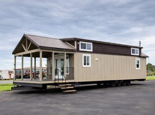A beige mobile tiny home with a sloped roof and small porch sits on an asphalt lot under a cloudy sky, conveying a sense of modern, compact living.